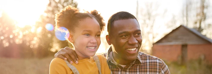 Chiropractic Service Area Chicago IL Dad and dotter A person and a child standing outdoors in a sunlit setting, with warm natural light creating a soft glow. The background includes a rustic wooden structure and leafless trees, suggesting a rural or countryside environment.